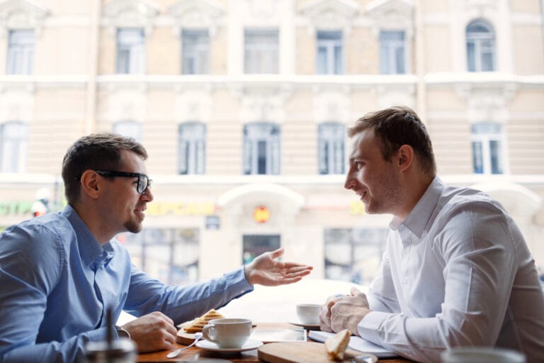 Zwei Männer in Geschäftskleidung unterhalten sich bei einer Tasse Kaffee an einem Cafétisch neben einem Fenster mit Blick auf die Straße der Stadt. Symbolisch für aktives Zuhören.