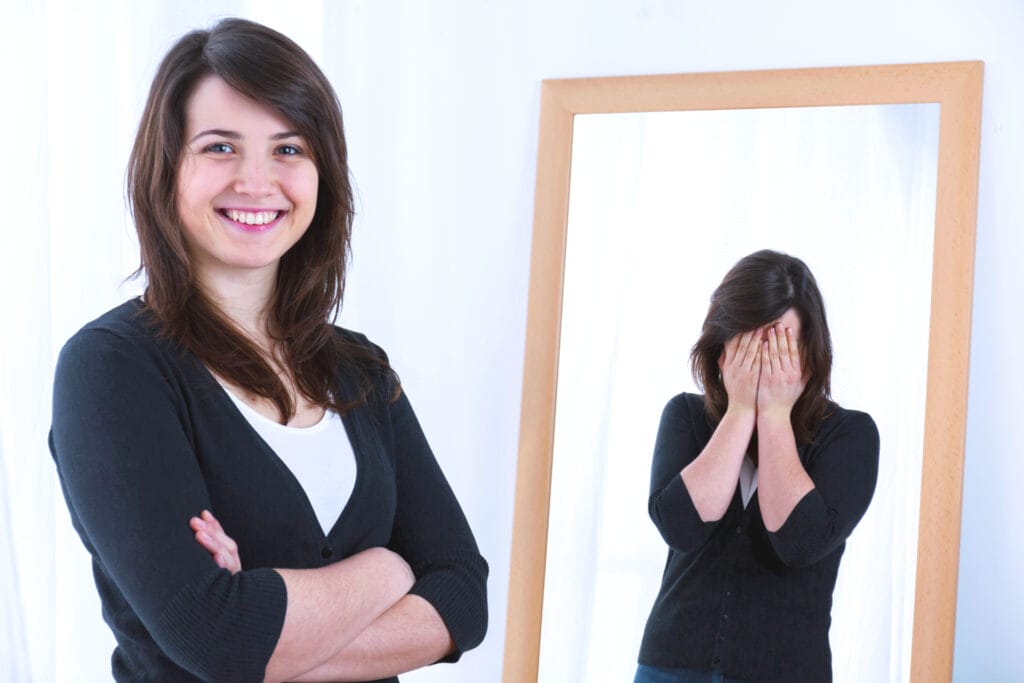 Eine junge Frau steht lächelnd vor einem Spiegel, während sich ihr Spiegelbild vor Trauer das Gesicht mit den Händen bedeckt. Symbolisches Bild für die Herausforderungen eines authentischen Lebensstils.