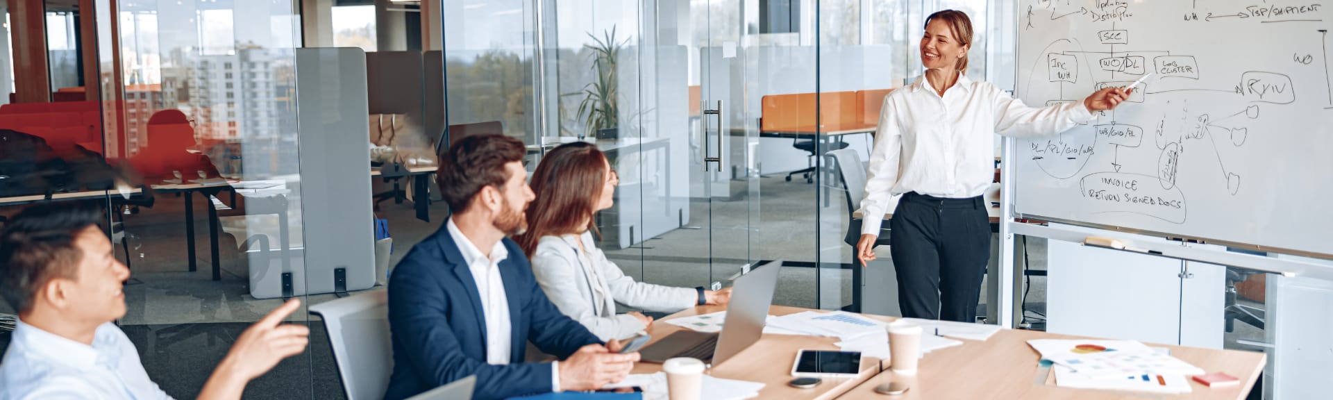 Eine Coachin steht in einem modernen Konferenzraum und erkl&auml;rt ihren Klienten ein Konzept am Whiteboard.