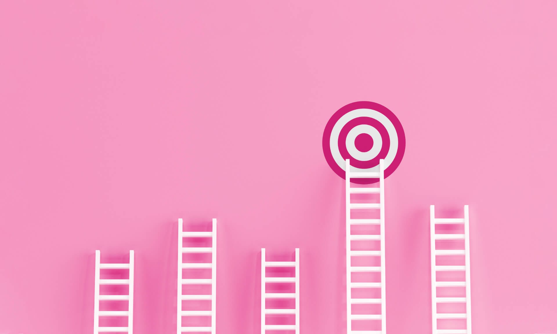 Eine Zielscheibe hängt an einer rosafarbenen Wand, die das Karriereziel symbolisiert. An dieser Wand sind fünf unterschiedlich große, weiße Leitern gelehnt, die Karriereleitern symbolisieren, aber nur eine Leiter führt zum Karriereziel.
