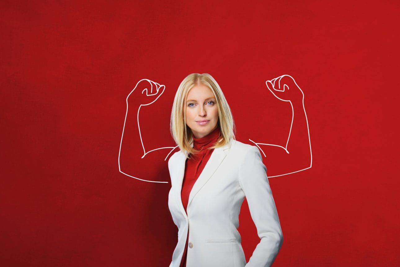 Eine Geschäftsfrau steht selbstbewusst im Raum. Auf der Wand hinter ihr sind muskulöse Arme aufgemahlt, die mit den Muskeln spielen. Sinnbild für verantwortungsbewusste, kompetente Mitarbeiter.