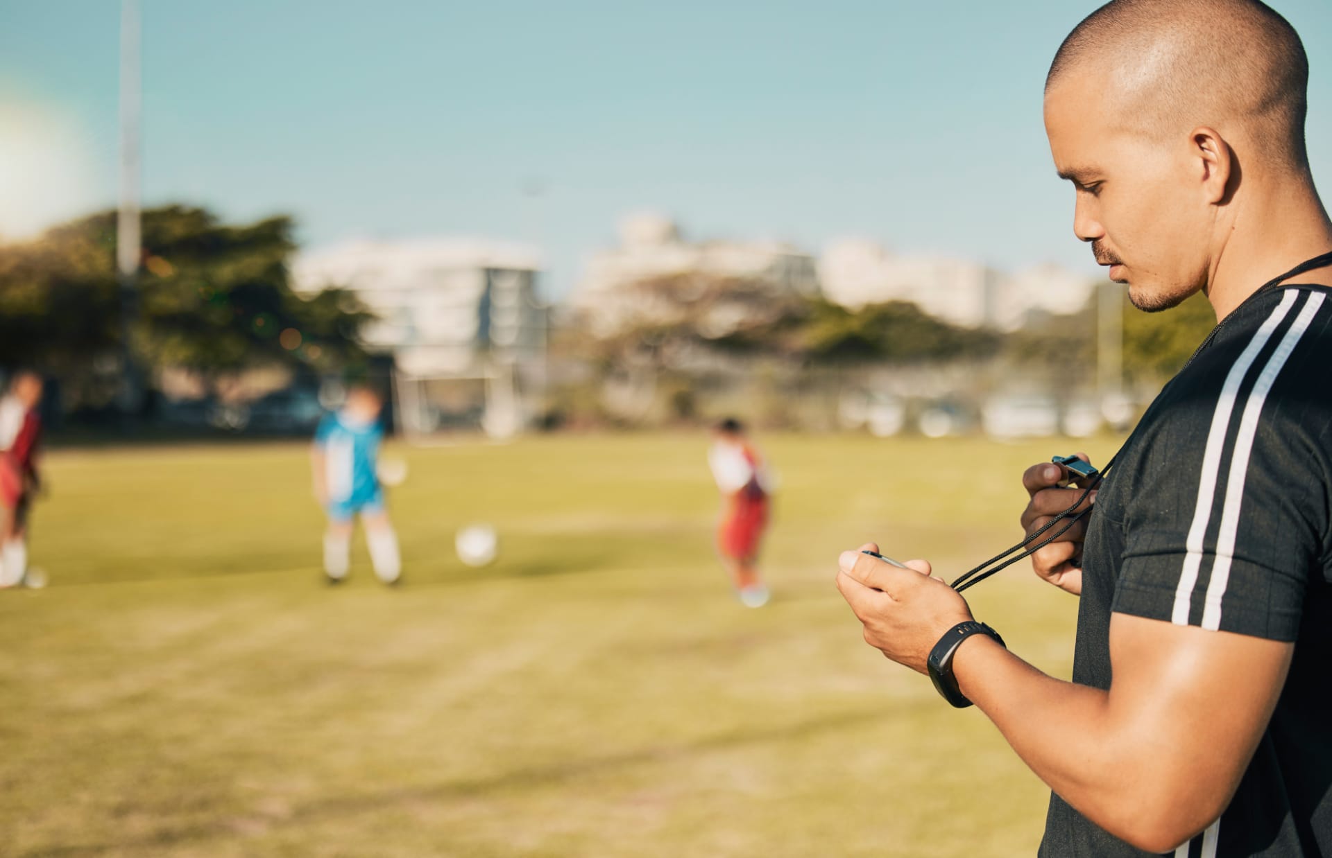 Ein professioneller Trainer benutzt auf einem Fu&szlig;ballfeld eine Stoppuhr, w&auml;hrend im Hintergrund die Spieler trainieren.