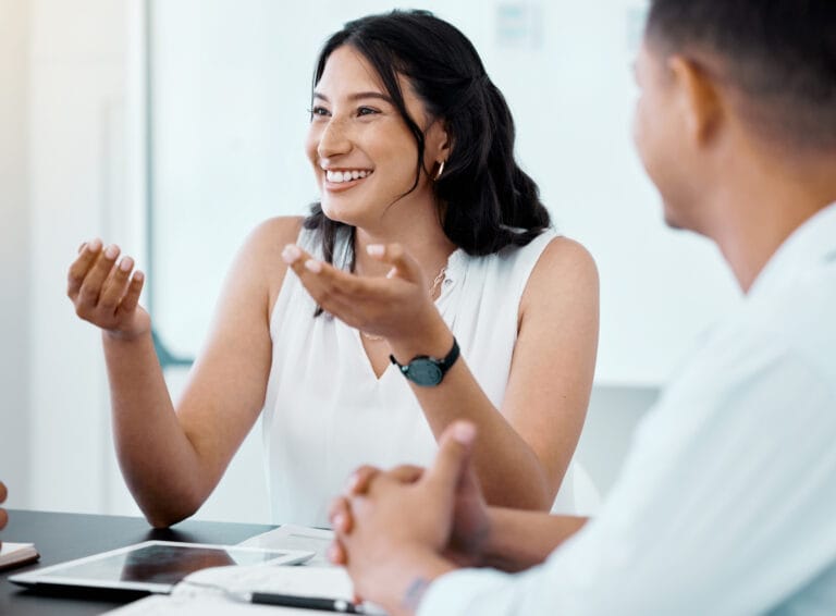 Eine Frau mit langen Haaren ist während einer Besprechung in eine Diskussion verwickelt, gestikuliert mit ihren Händen und lächelt, wobei sie wahrscheinlich Techniken aus der Transaktionsanalyse anwendet, während eine andere Person aufmerksam zuhört.