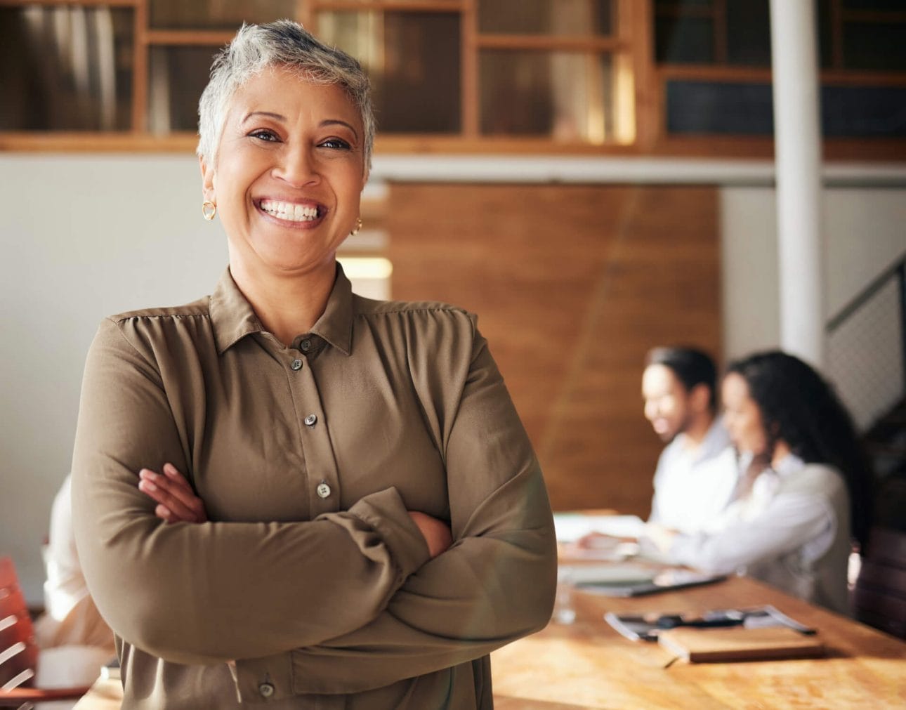 Eine Verantwortungsträgerin steht mit einer selbstbewussten Körperhaltung lächelnd in einem Büro, während ihre Mitarbeiterinnen und Mitarbeiter im Hintergrund am Meetingtisch arbeiten