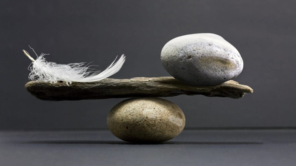 Auf der einen Seite einer Waage liegt eine Feder, auf der anderen ein Stein. Die Waage ist im Gleichgewicht. Symbolisches Bild für die Balance aus Feingefühl und Autorität in der Personalführung.