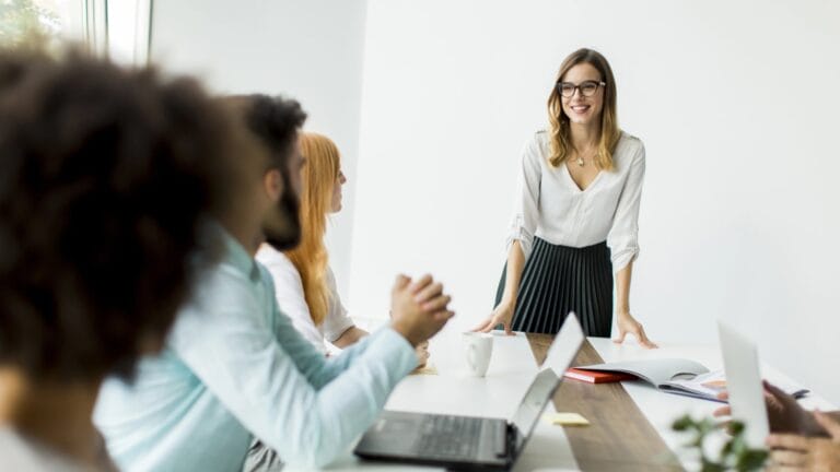 Eine junge Teamleiterin steht im Meetingraum und spricht mit einem freundlichen Gesichtsausdruck und Autorität vor ihren Mitarbeitern. Symbolisches Bild, wie man sensibel und doch kraftvoll führen kann.