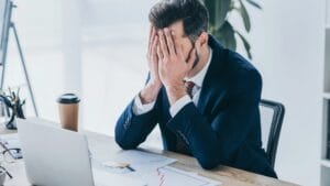 Ein Mann sitzt am Arbeitsplatz und bedeckt vor Scham sein Gesicht mit den Händen. Symbolisches Bild für Scham im Beruf.