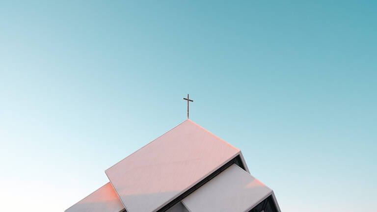 Das Kreuz auf der Dachspitze einer Kirche ragt in den blauen Himmel. Symbolisch für karitative Dienste und diakonisch-kirchliche Einrichtungen.