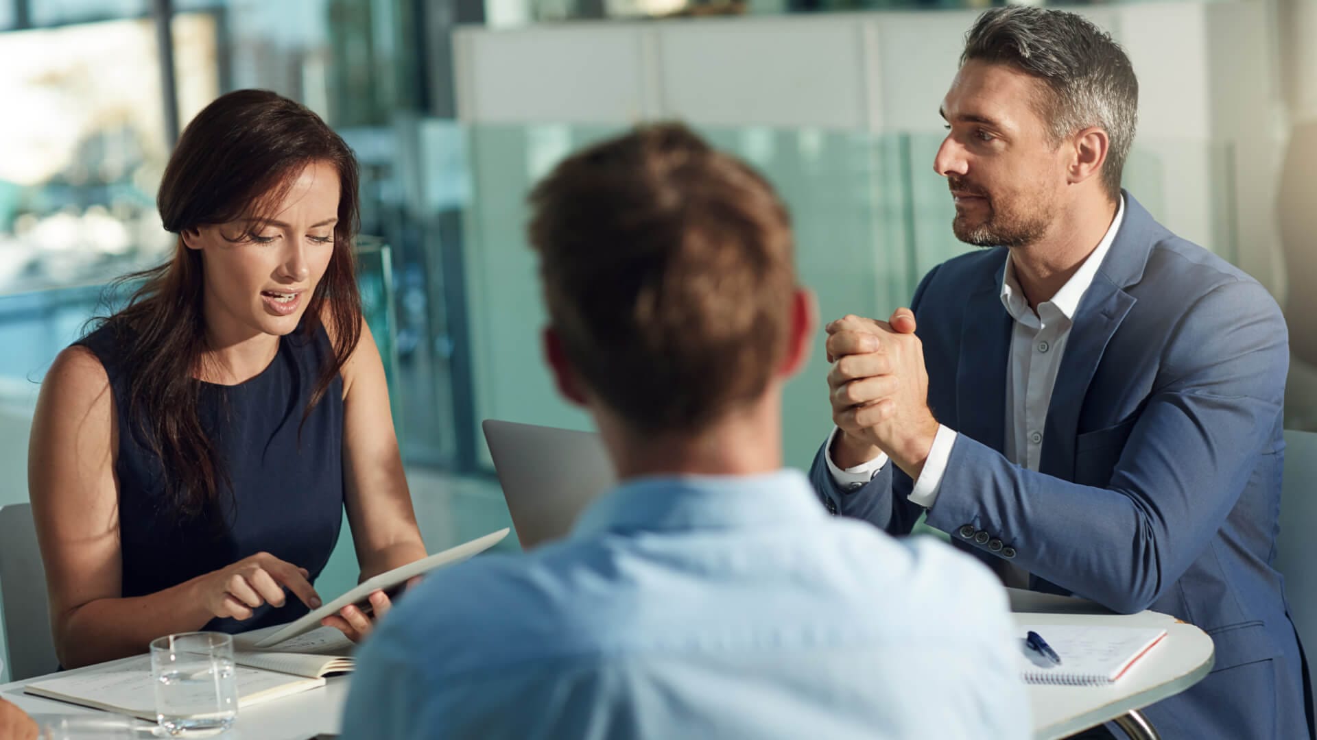 Ein Team sitzt am Besprechungstisch. Der Leiter hört einer Kollegin im Rahmen eines Teamtrainings zu.
