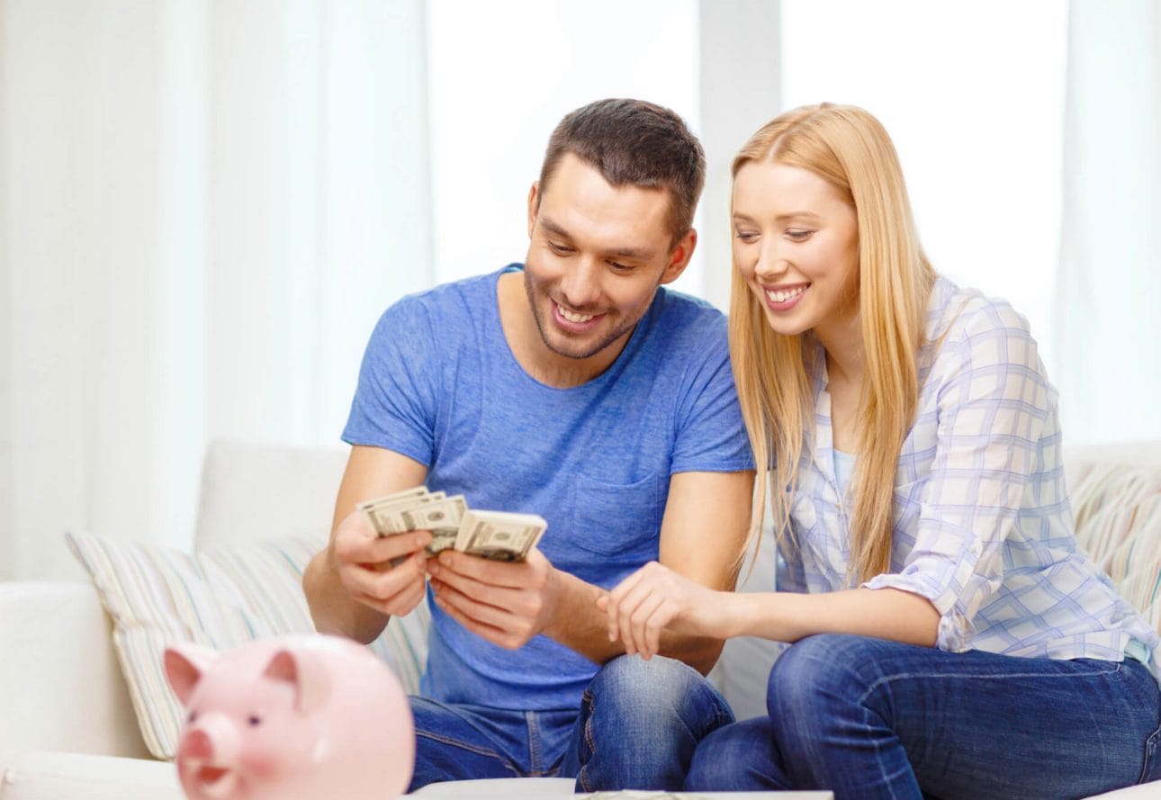 Ein Mann und eine Frau sitzen auf einem Sofa und zählen einen Bündel Geld, während ein Sparschwein auf einem Tisch vor ihnen steht. Symbolisch für die gesparte Summe durch steuerfreie Ausbildungen.