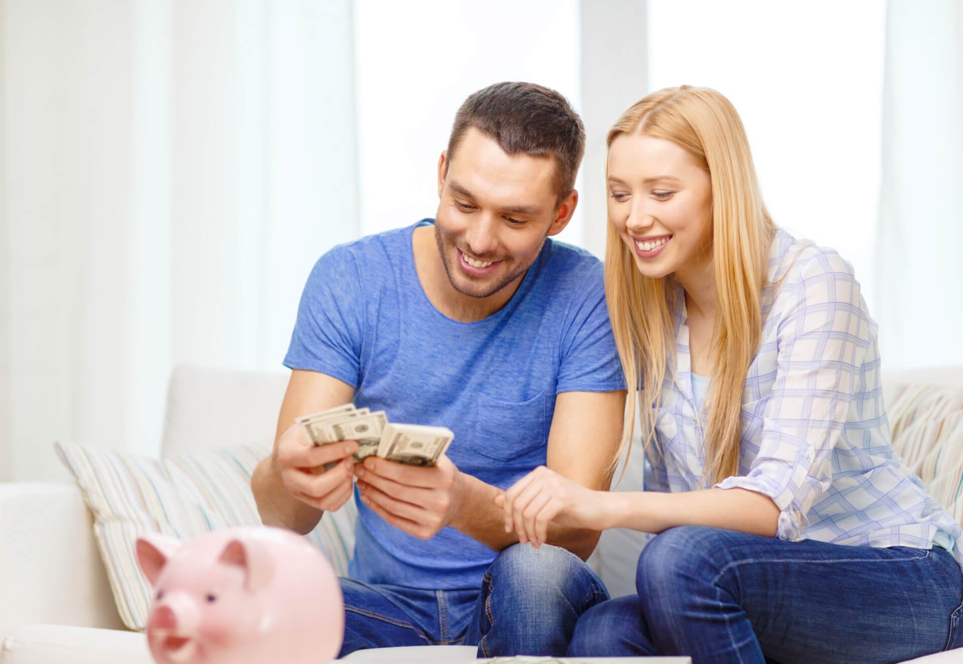 Ein Mann und eine Frau sitzen auf einem Sofa und zählen einen Bündel Geld, während ein Sparschwein auf einem Tisch vor ihnen steht. Symbolisch für die gesparte Summe durch steuerfreie Ausbildungen.