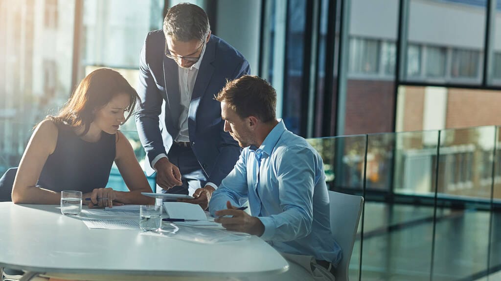 Ein Mann und eine Frau sitzen am Tisch im Meetingraum, während ein Supervisor neben ihnen steht und am Tablet die Ergebnisse ihrer Arbeit verfolgt. Symbolisches Bild für eine Supervision in Nürnberg, Fürth, Erlangen und Umgebung.